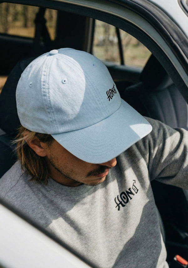 Homme portant une casquette HONO bleu clair et un sweat gris brodé HONO, assis dans une voiture, esprit surfwear durable et lifestyle naturel