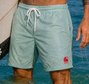 Boardshort homme recyclé HONO vert d'eau porté dans l'océan avec une planche de surf, surfwear durable