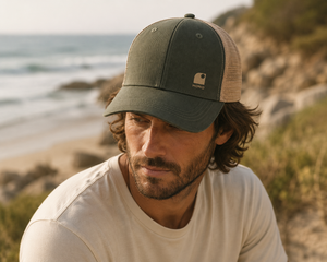 Homme portant une casquette trucker HONO kaki et beige avec logo brodé beige, photographié en bord de mer sur un sentier côtier. Casquette surfwear éco-responsable en coton biologique et polyester recyclé, style premium inspiré de l'univers du surf.