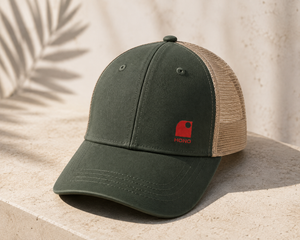 Casquette trucker HONO kaki et beige avec logo corail, présentée sur une pierre naturelle dans une ambiance lumineuse inspirée du bord de mer, style surfwear premium.