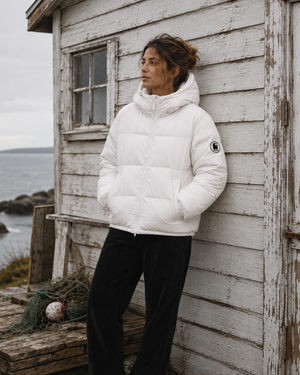 Doudoune blanche chaude en matières recyclées HONO, style surfwear, portée par une femme devant une cabane de pêcheur avec du bois et un poêle à bois.
