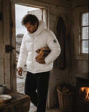 Doudoune blanche chaude en matières recyclées HONO, style surfwear, portée par un homme entrant dans une cabane de pêcheur avec du bois et un poêle à bois.