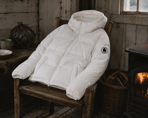 Doudoune blanche chaude en matières recyclées HONO, style surfwear, mise en scène dans une cabane de pêcheur avec poêle à bois et ambiance hivernale.