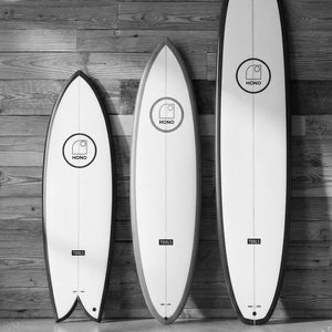 Trois planches de surf HONO en noir et blanc posées contre un mur en bois dans l’atelier du shaper, représentant différents shapes du fish au mini long, illustrant le travail artisanal et le design des boards.