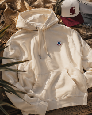Hoodie à capuche HONO coton bio écru, sweat surf éco-responsable avec logo poitrine, ambiance surf lifestyle sur terrasse en bois au bord de l'océan.