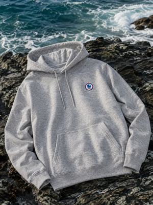 Hoodie à capuche HONO coton bio gris chiné, sweat surf éco-responsable avec logo poitrine sur des rochers en bord d'océan.