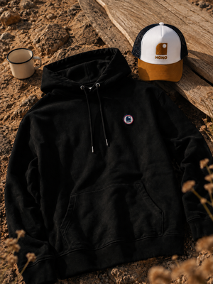 Hoodie à capuche HONO coton bio noir, sweat surf éco-responsable avec logo poitrine, mise en scène plage, sable et accessoires surf lifestyle.