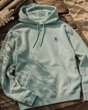 Hoodie à capuche HONO  Surf en coton bio vert d'eau avec logo poitrine, sweat surf éco-responsable posé sur une terrasse en bois face à la plage, ambiance surf lifestyle.