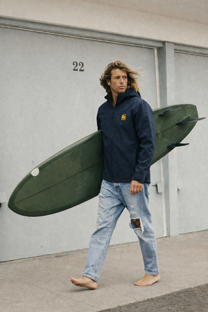 Homme marchant avec planche de surf portant veste softshell hiver outdoor HONO marine recyclée 3 couches