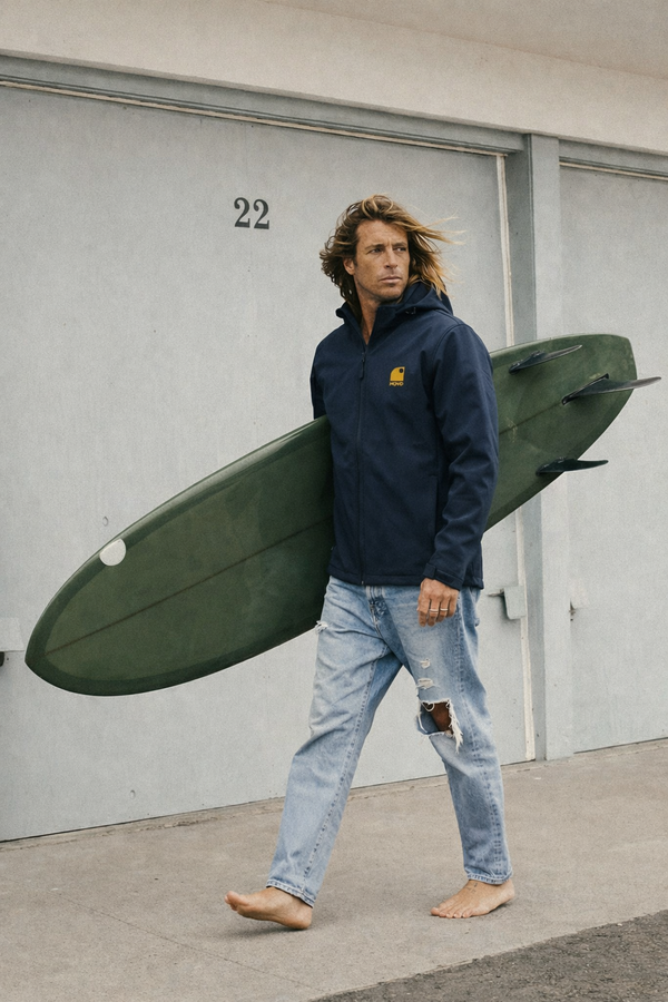 Homme marchant avec planche de surf portant veste softshell hiver outdoor HONO marine recyclée 3 couches