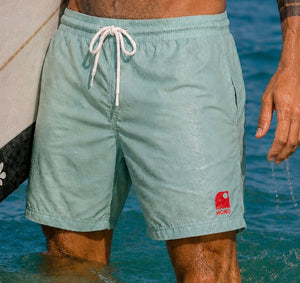 Boardshort homme recyclé HONO vert d'eau porté dans l'océan avec une planche de surf, surfwear durable