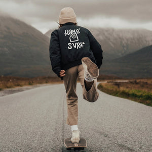 Lifestyle veste bomber noir durable Surf HONO Signature TIKI portée en skateboard dans un paysage montagneux