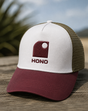 Casquette trucker recyclée HONO avec logo brodé bordeaux, panneau avant blanc, visière bordeaux et arrière en mesh kaki recyclé, photographiée en extérieur sur une table en bois.