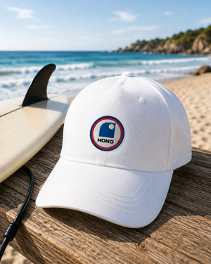 Casquette surf blanche recyclée HONO avec pbroderie tricolore posée sur une table en bois face à l'océan, à côté d'une planche de surf, dans une ambiance bord de mer lumineuse