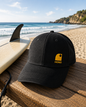 Casquette surf noire recyclée HONO avec broderie jaune posée sur une table en bois face à l'océan, à côté d'une planche de surf, dans une ambiance bord de mer lumineuse