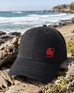 Casquette surf noire HONO Wave broderie corail en coton recyclé posée sur du bois flotté face à l'océan, ambiance surfwear premium