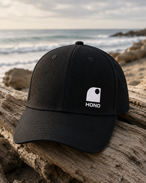 Casquette surf recyclée HONO Wave noire avec logo brodé blanc, photographiée sur du bois flotté face à l'océan dans une ambiance bord de mer