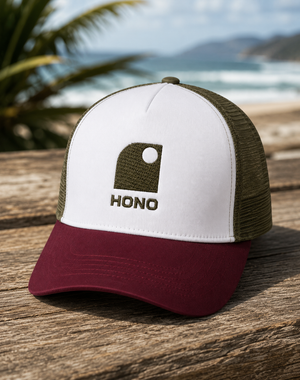 Casquette surf trucker recyclée HONO avec logo brodé kaki, panneau avant blanc, visière bordeaux et arrière en mesh kaki recyclé, photographiée en extérieur sur une table en bois face à l'océan.