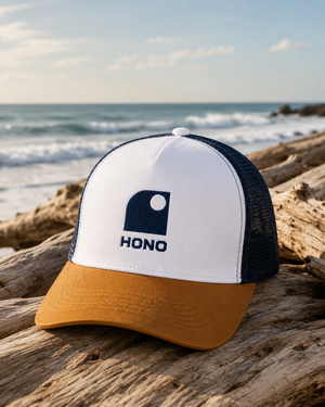 Casquette surf trucker recyclée HONO avec logo brodé navy, panneau avant blanc, visière camel et arrière en mesh navy recyclé, photographiée sur du bois flotté face à l'océan