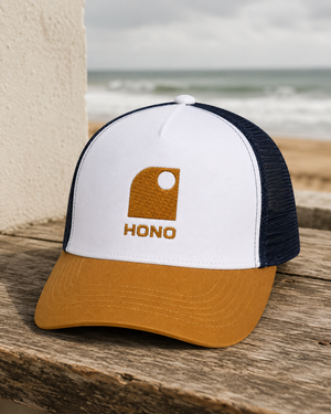 Casquette surf trucker recyclée HONO avec logo brodé camel, panneau avant blanc, visière camel et arrière en mesh navy recyclé, photographiée sur une table en bois face à l'océan.