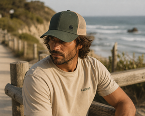 Homme portant une casquette trucker HONO kaki avec filet beige et logo Wave brodé noir, photographié sur un sentier côtier face à l'océan dans une ambiance surf lifestyle naturelle.