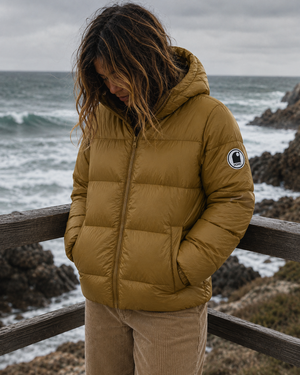 Doudoune chaude en nylon recyclé HONO Surfwear bronze portée par une femme sur la côte, face à l’océan