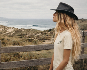 femme surfeuse portant un t-shirt beige HONO surfwear avec logo palmier style surf lifestyle au bord de l ocean 