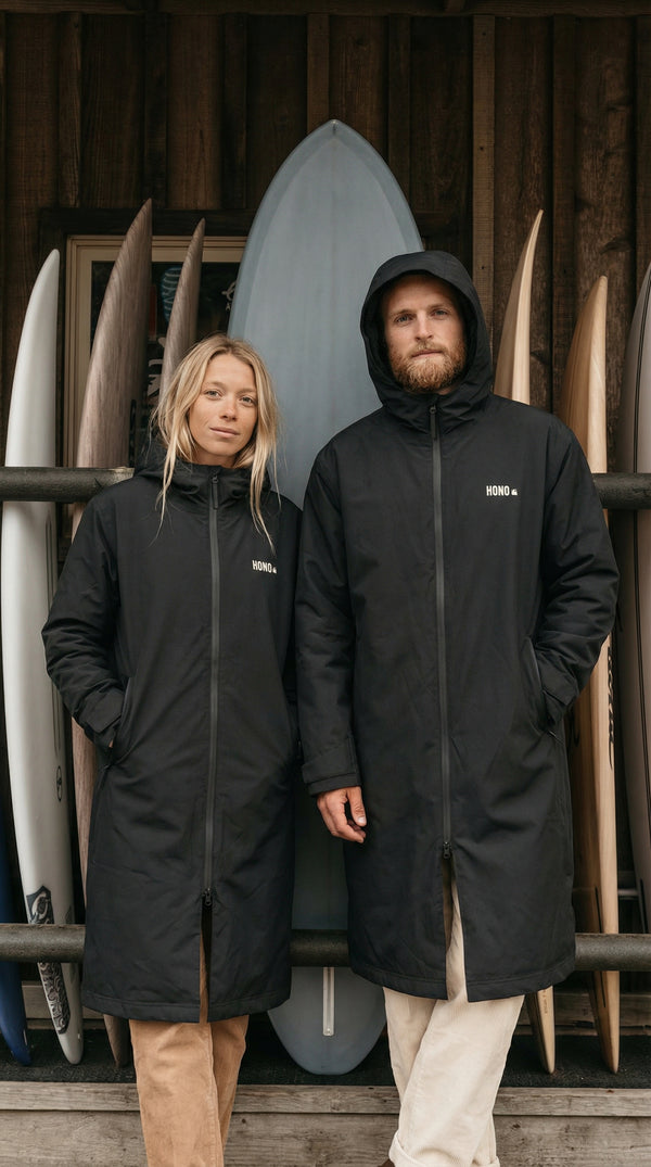 Parka longue imperméable écoresponsable HONO noire, modèle technique unisexe surf durable, portée devant des planches de surf