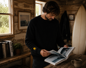 Pull HONO Surf Coastlife homme noir en coton bio et polyester recyclé porté dans une cabane en bois avec un livre de surf