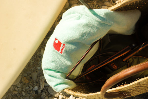 Détail broderie short éponge femme HONO surfwear durable dans un sac de plage près d’une planche de surf