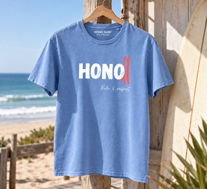 T-shirt HONO Surf bleu en coton bio au style vintage, photographié sur la plage avec une planche de surf