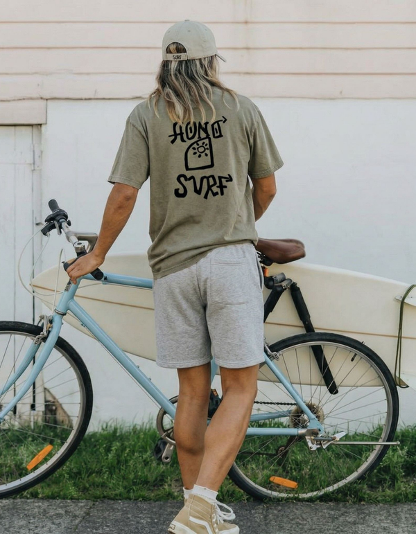 Homme de dos portant le t-shirt surf HONO Tiki kaki vintage en coton bio avec logo HONO SURF noir, appuyé sur un vélo