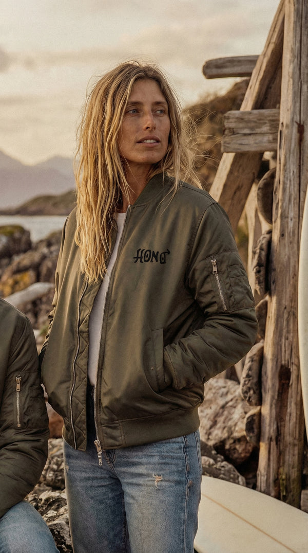 Veste bomber kaki HONO Surf Signature TIKI portée par une femme dans un décor côtier avec planche de surf